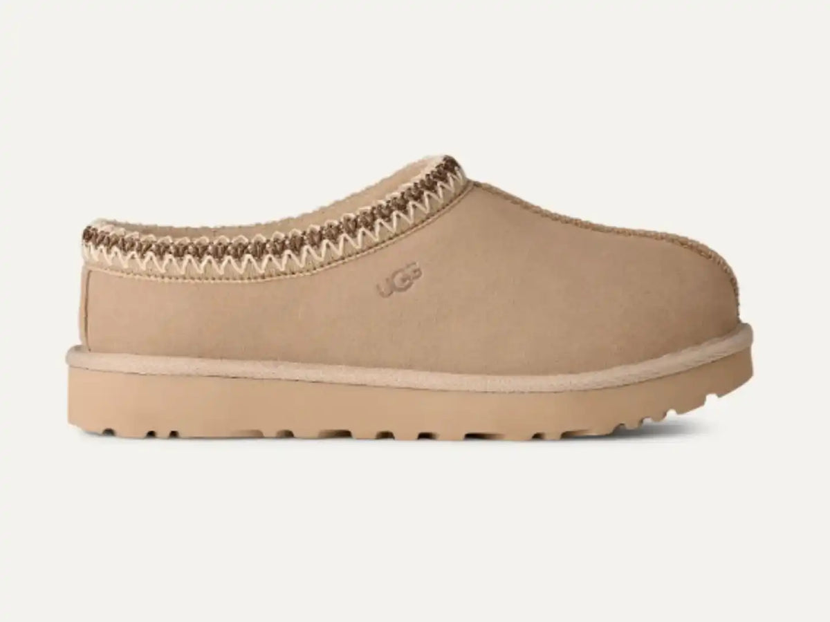 A single beige suede slip-on clog shoe with a braided trim detail and a thick lug sole, shown in profile against a light off-white background. The UGG logo is subtly embossed on the side.