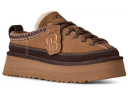 A brown suede and dark brown ribbed knit platform sneaker by UGG, featuring a thick tan sole, dark brown laces, a tan leather UGG logo patch on the side, and a white faux-fur collar, shown against a plain white background.