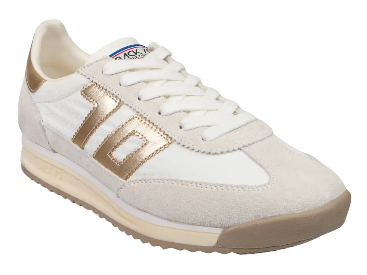 A low-top sneaker in off-white suede and leather with metallic gold accents on the side stripe and heel tab, white laces, and a beige midsole and outsole.