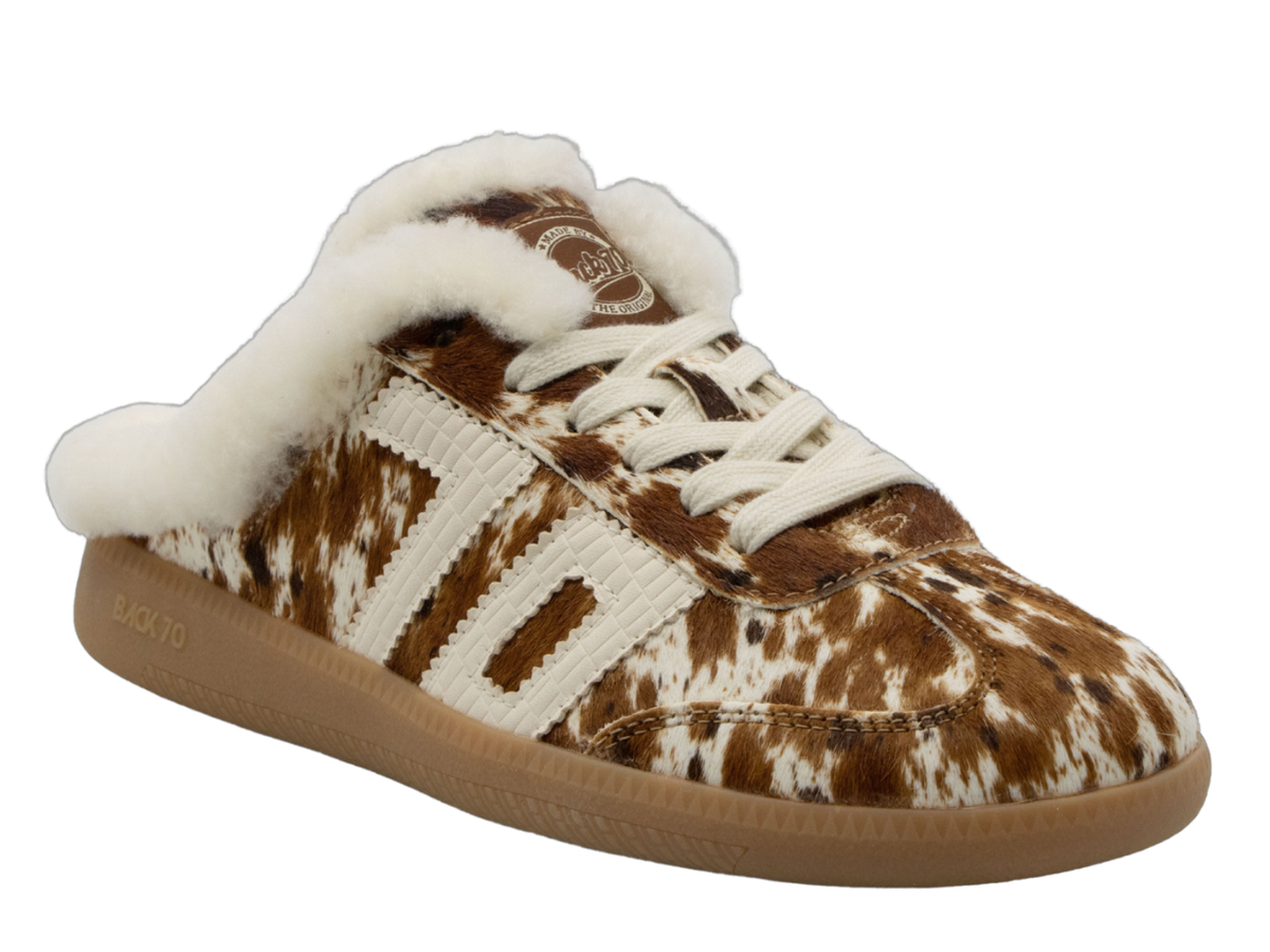 A low-top slip-on sneaker featuring a brown and white cowhide pattern, cream knit side stripes, a thick white faux-fur collar, and a brown gum sole.