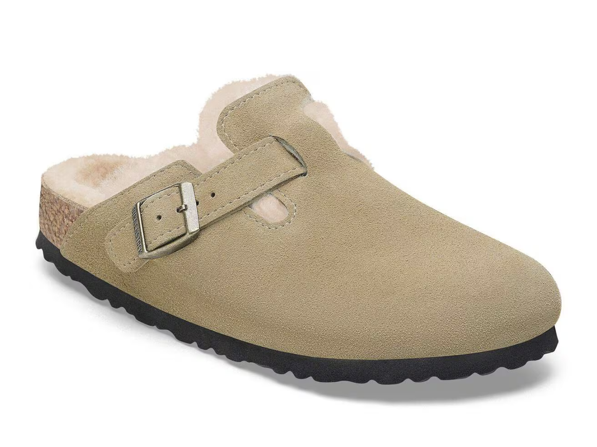 A single taupe suede clog-style slipper with a black sole and a light beige sherpa lining, featuring a single adjustable strap with a silver buckle.