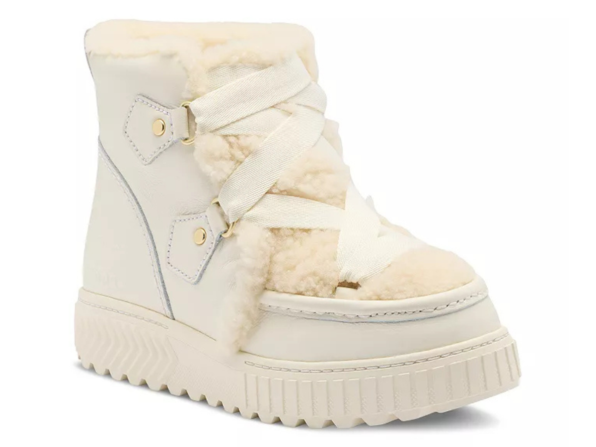 A cream-colored platform ankle boot with a plush faux-shearling lining, wide decorative straps, and gold-tone hardware, shown against a plain white background.